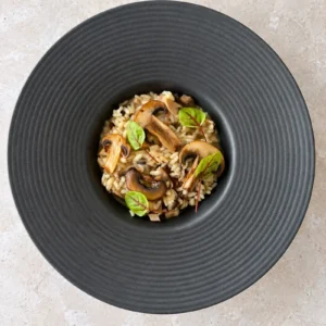 Overhead view of creamy vegan mushroom risotto served in a dark wide rim bowl, topped with golden mushroom slices and fresh microgreens.