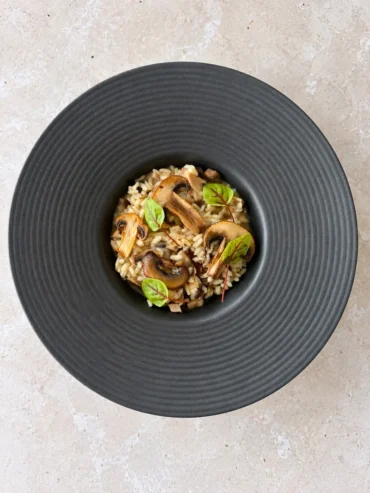 Overhead view of creamy vegan mushroom risotto served in a dark wide rim bowl, topped with golden mushroom slices and fresh microgreens.