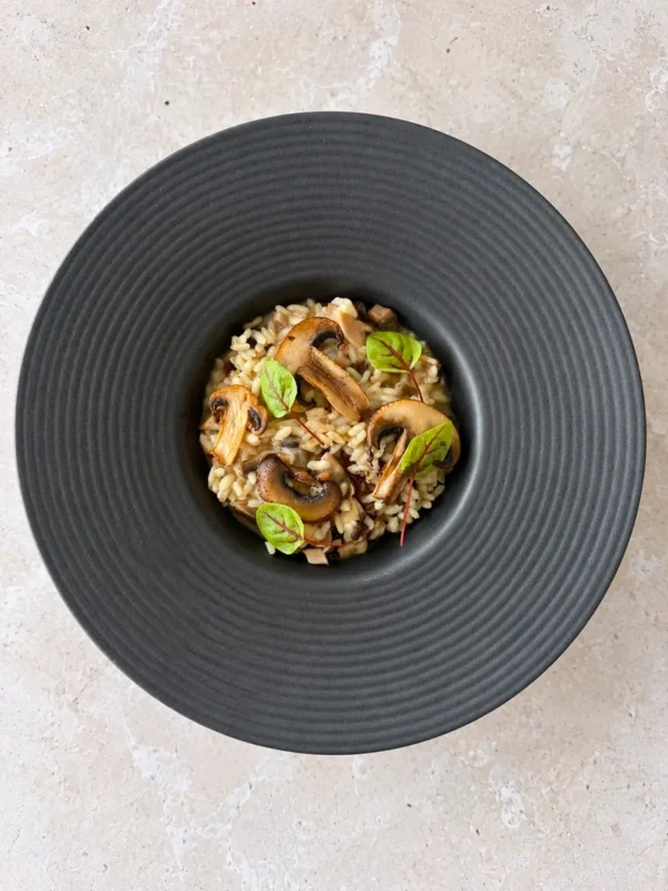 Overhead view of creamy vegan mushroom risotto served in a dark wide rim bowl, topped with golden mushroom slices and fresh microgreens.