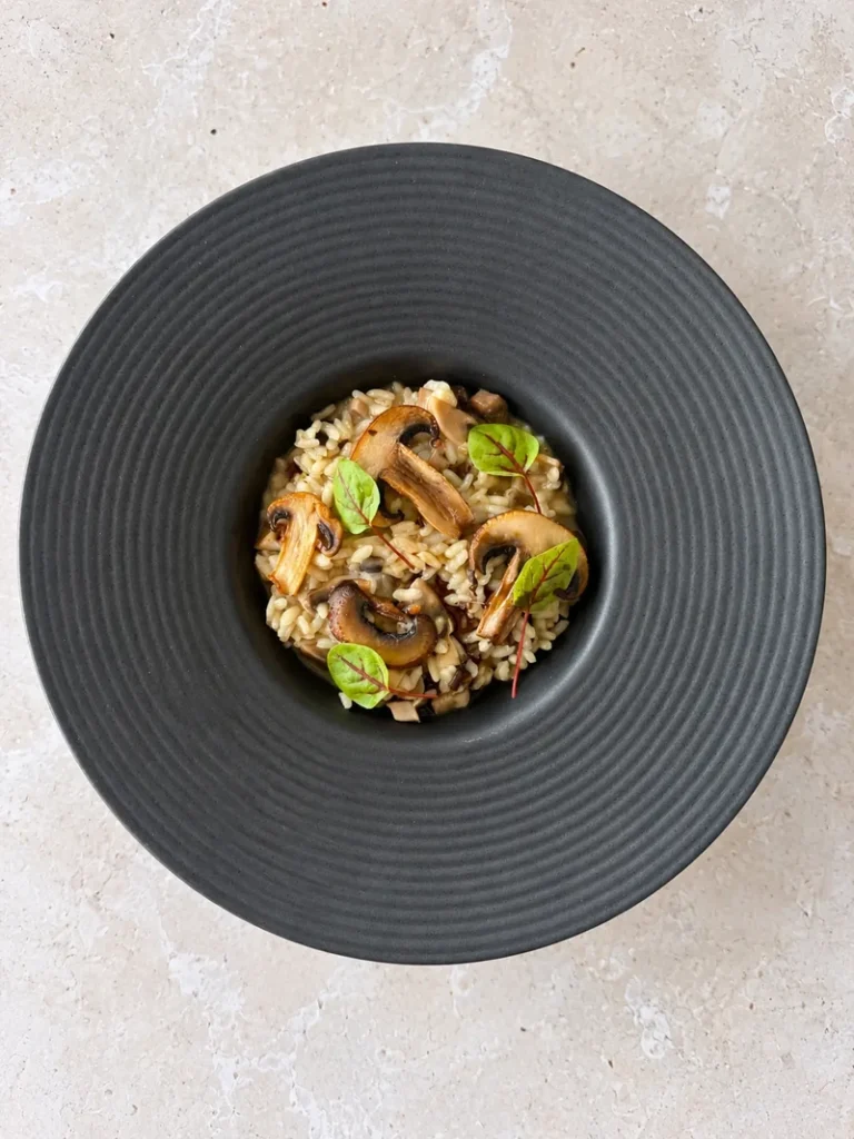 Overhead view of creamy vegan mushroom risotto served in a dark wide rim bowl, topped with golden mushroom slices and fresh microgreens.