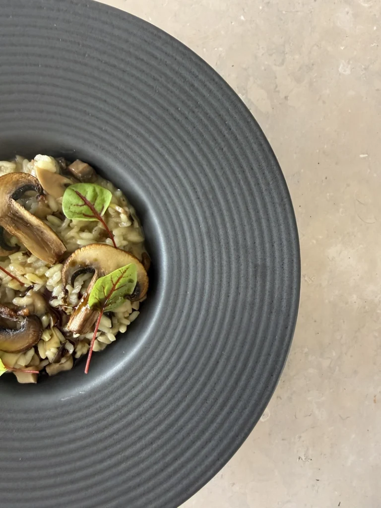 Close up of creamy vegan mushroom risotto topped with golden mushrooms and fresh microgreens, served in a dark ceramic bowl.