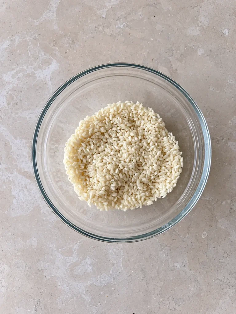 Uncooked risotto rice in a glass bowl on a light stone surface, ready for vegan mushroom risotto.