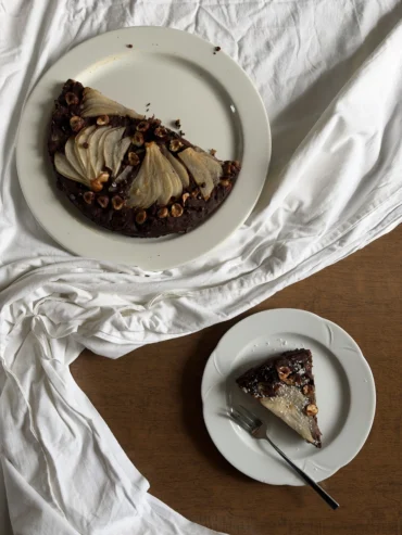 Vegan chocolate pear cake with sliced pears, toasted hazelnuts and desiccated coconut, served with an individual slice on a white plate.