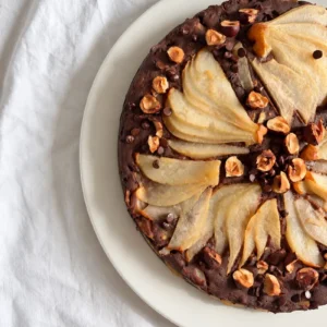 Vegan chocolate pear cake topped with sliced pears, toasted hazelnuts and dark chocolate chips on a white plate.