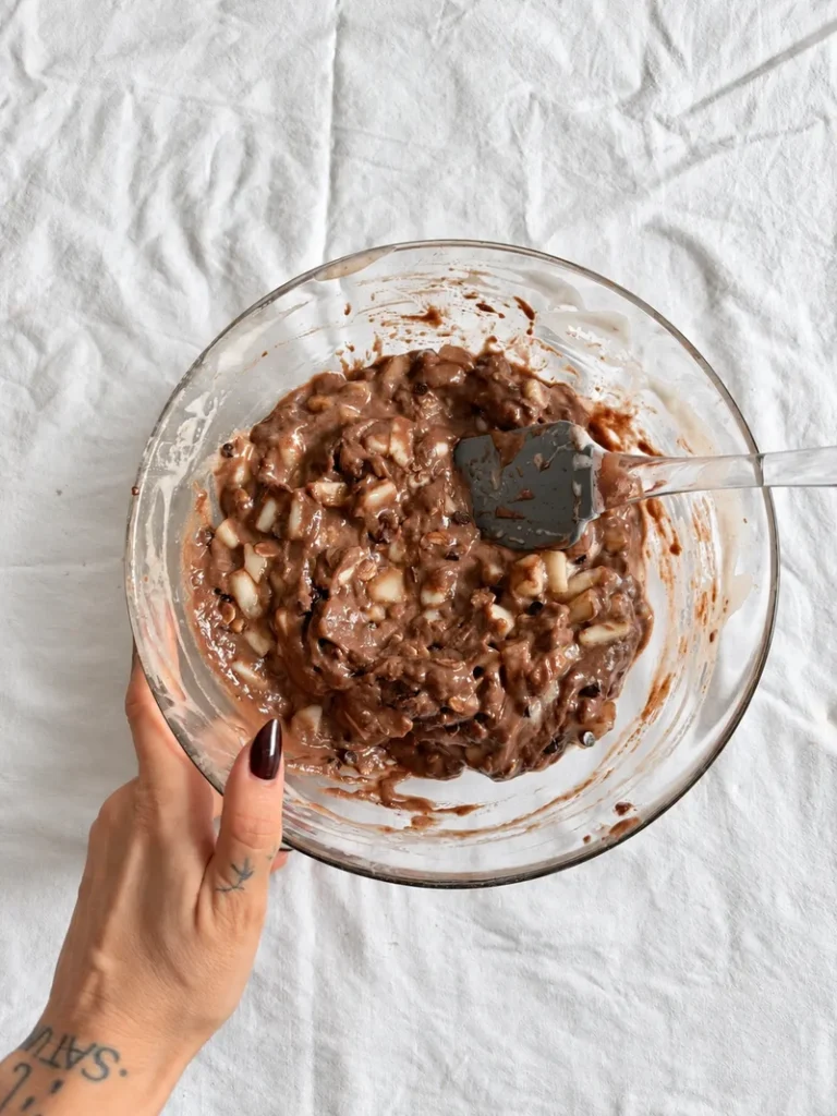 Wet and dry ingredients for vegan chocolate pear cake in a glass bowl before mixing, with cocoa powder and chocolate pieces visible.