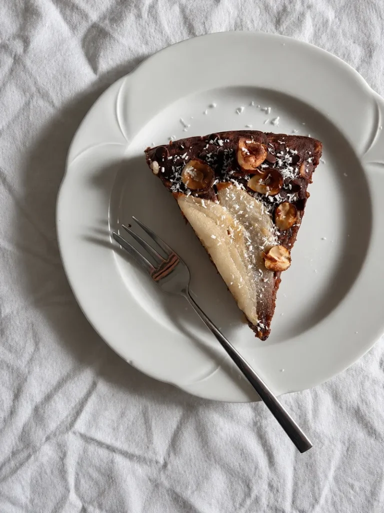 Slice of vegan chocolate pear cake topped with hazelnuts and desiccated coconut, served on a white plate with a fork.