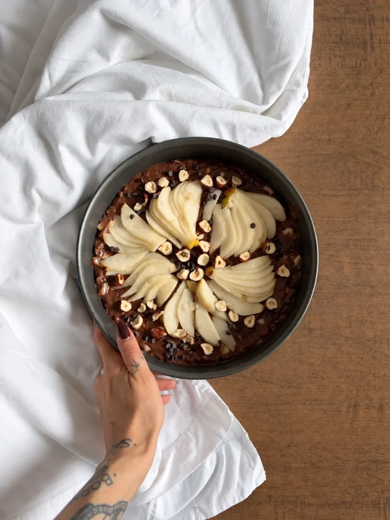 Vegan chocolate pear cake before baking, topped with sliced pears, chopped hazelnuts and dark chocolate chips in a round cake tin.