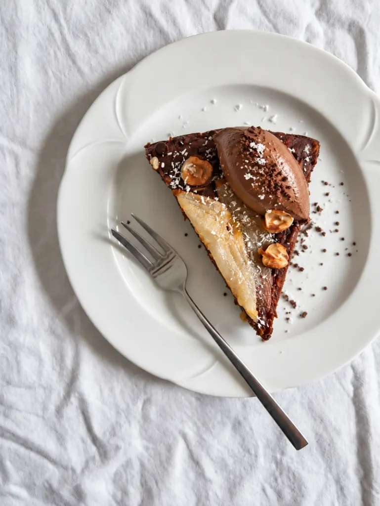 Slice of vegan chocolate pear cake topped with a quenelle of vegan chocolate mousse, hazelnuts and desiccated coconut.