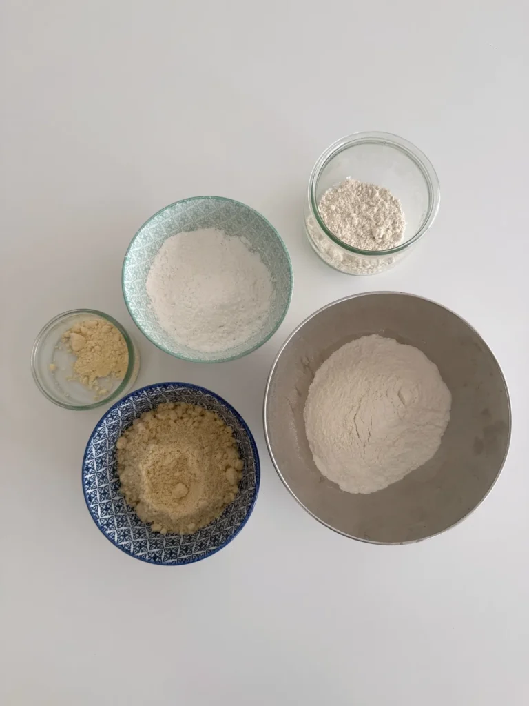 Different gluten-free flours arranged in bowls on a white surface, ready to prepare homemade healthy apple bread.