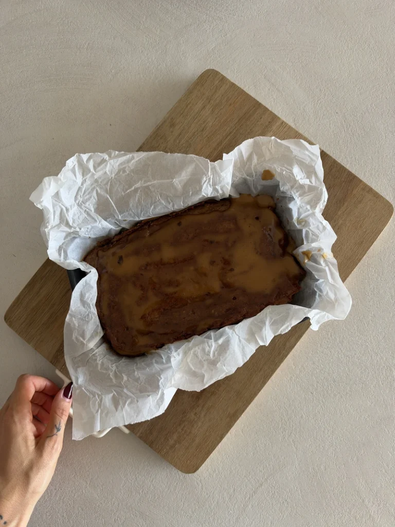 Overhead view of a freshly baked vegan gluten free sticky toffee pudding in a parchment lined baking dish, topped with warm miso toffee sauce and resting on a wooden board.