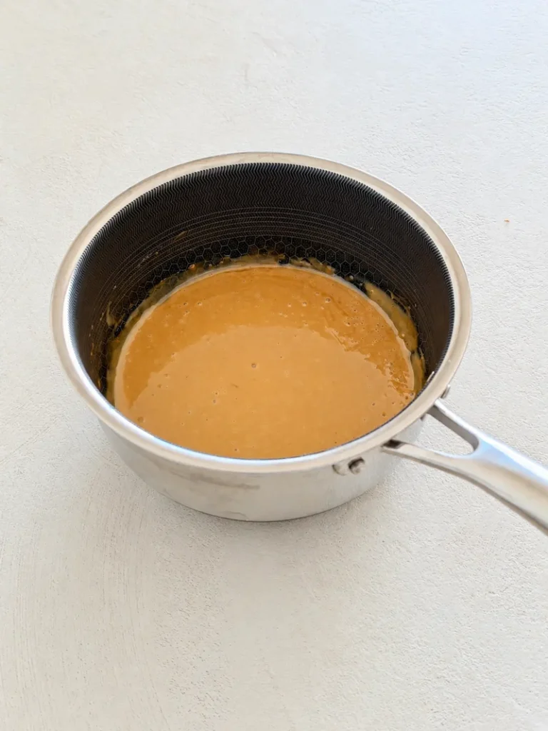 Vegan miso toffee sauce in a small saucepan, smooth and glossy, photographed on a light textured surface.