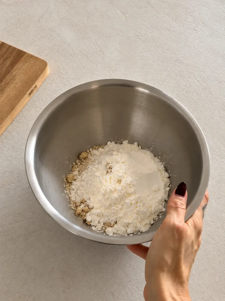 Hand holding a stainless steel mixing bowl filled with the dry ingredients for vegan gluten free sticky toffee pudding on a light textured countertop.