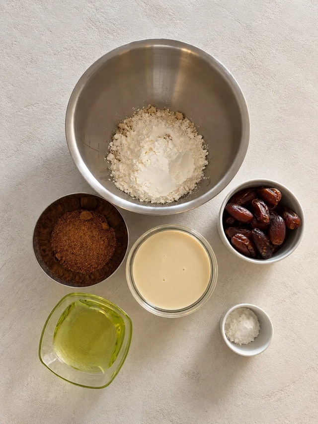 Overhead view of the ingredients for vegan sticky toffee pudding arranged on a light textured surface, including a bowl of flour, Medjool dates, coconut sugar, soy milk, oil, and a small bowl of baking powder.