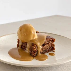 Slice of sticky toffee pudding topped with a scoop of plant based ice cream and covered in glossy miso toffee sauce on a cream plate.