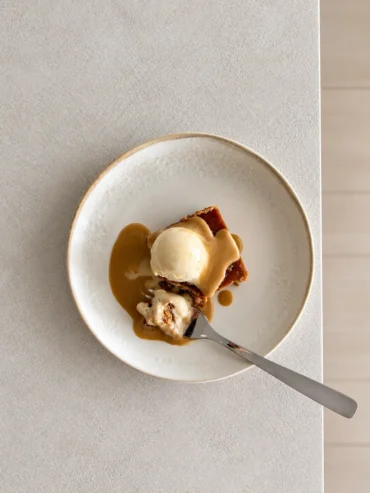 Overhead view of a plated serving of sticky toffee pudding topped with plant based vanilla ice cream and glossy miso toffee sauce, with a fork taking a bite from the side.