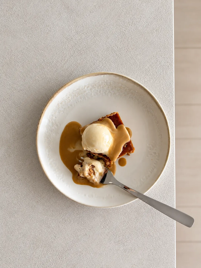 Overhead view of a plated serving of sticky toffee pudding topped with plant based vanilla ice cream and glossy miso toffee sauce, with a fork taking a bite from the side.